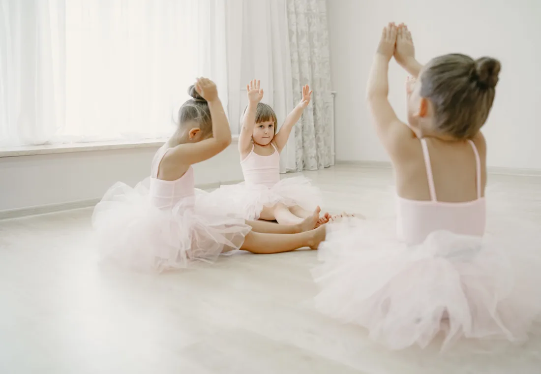Drie kleine ballerina's in tutu zitten met gestrekte armen op de studiovloer